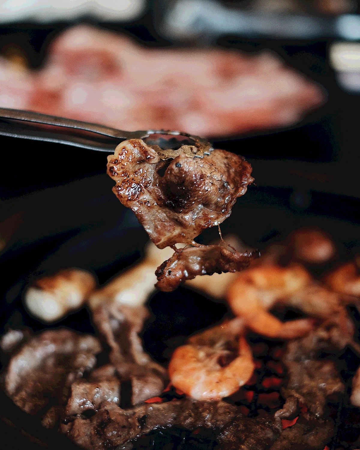 Meat cooking on a Korean BBQ, with a done piece of beef held by tongs