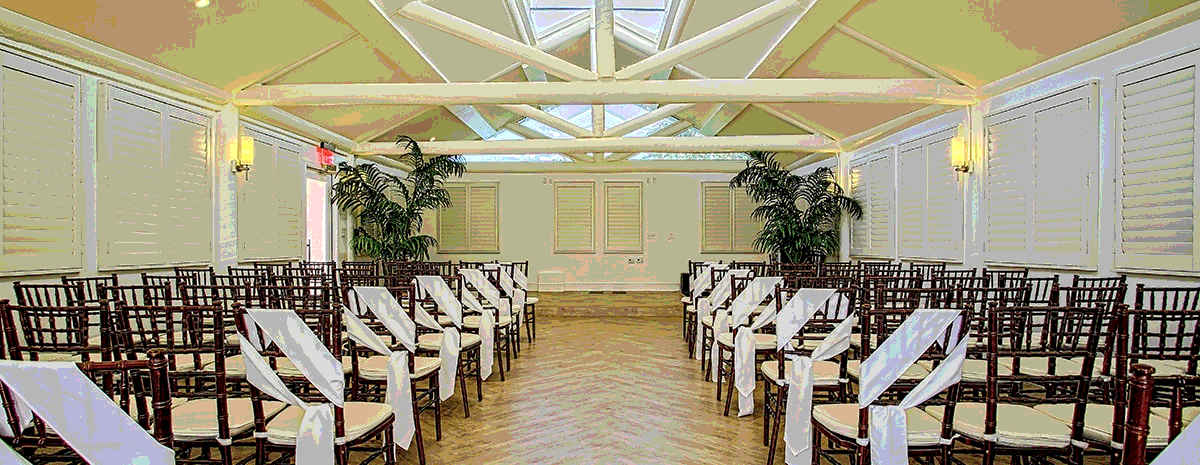 A photo facing the dais of the wedding chapel, in a tropical island theme with wooden slatted shutters, dark wood chairs, exposed beams, in whites and creams